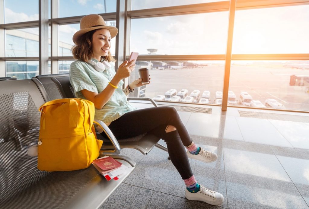 jovem sentada em um aeroporto esperando pelo voo. Ela está sorrindo e mexendo no celular