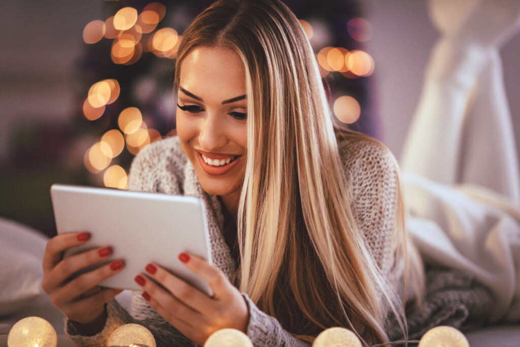 Jovem bonita sorridente usando tablet para vender milhas e sorrindo feliz durante as férias de Natal aconchegantes em casa.