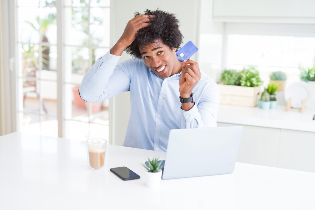 Homem afro-americano usando laptop e compras online com cartão de crédito estressado com a mão na cabeça, intrigado sobre os mitos dos programas de milhagem.