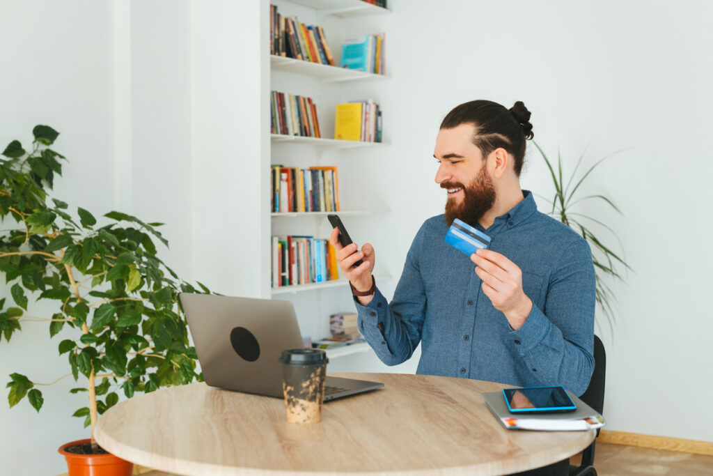 Homem alegre trabalhando no escritório e usando cartão de crédito e smartphone.