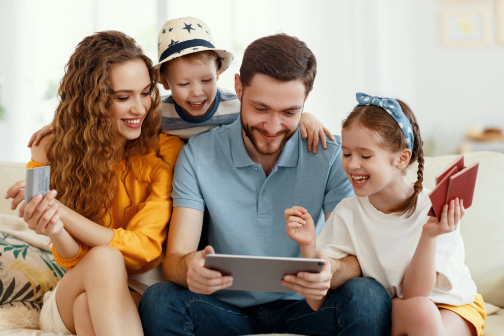 Pais sorridentes com crianças reunidas no sofá usando tablet enquanto escolhem uma excursão on-line antecipando as férias de verão todos juntos. Eles participam do programa de pontos e milhas qualificáveis.