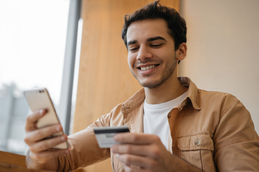 Um homem de camis amarrom está segurando um celular em uma mão e um cartão de crédito na outra, ele sorri para o celular enquanto acumula pontos C6 Bank com sua compra