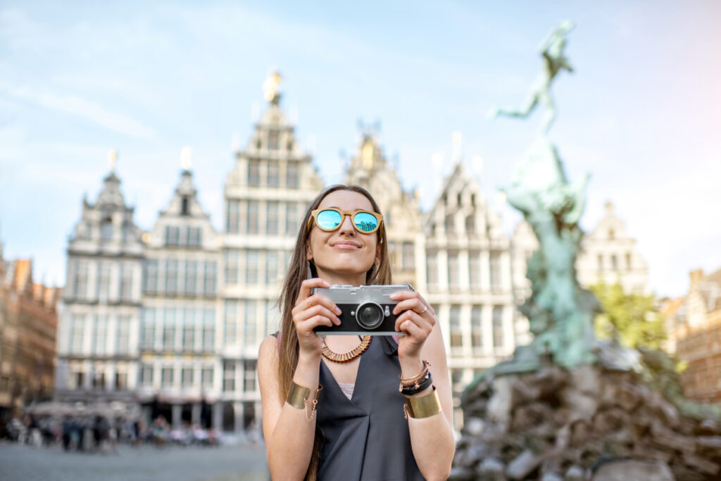 Uma mulher branca, de cabelo liso, óculos de sol com lente espelhada, blusa cinza e pulseiras está turistando pela Europa. Ela segura uma camera fotográfica e sorri enquanto registra o seu mochilão na Europa