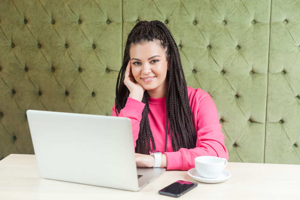 Retrato de uma linda jovem satisfeita com penteado de dreadlocks preto na blusa rosa, sentado no café, trabalhando no laptop com um sorriso, segurando uma mão no queixo, olhando para a câmera.