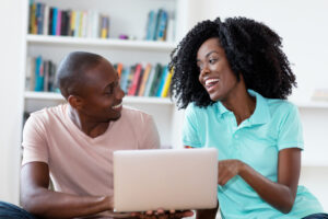 Casal afro-americano rindo com o computador dentro de casa avaliando formas de ganhar milhas.