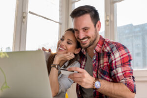Jovem casal feliz realizando compras em seu notebook com cartão de crédito para acumular milhas e aproveitar a promoção de milhas aéreas e ter lucros.
