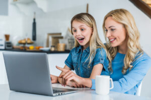 Mãe sorridente com filha usando laptop na cozinha.