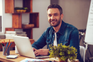 Jovem confiante trabalhando em um laptop e sorrindo enquanto está sentado em seu local de trabalho no escritório.