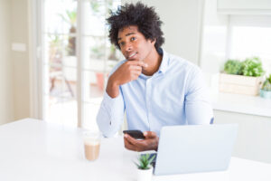 Homem de negócios afro-americano usando smartphone e laptop rosto sério pensando na pergunta sobre mitos dos programas de milhagem.