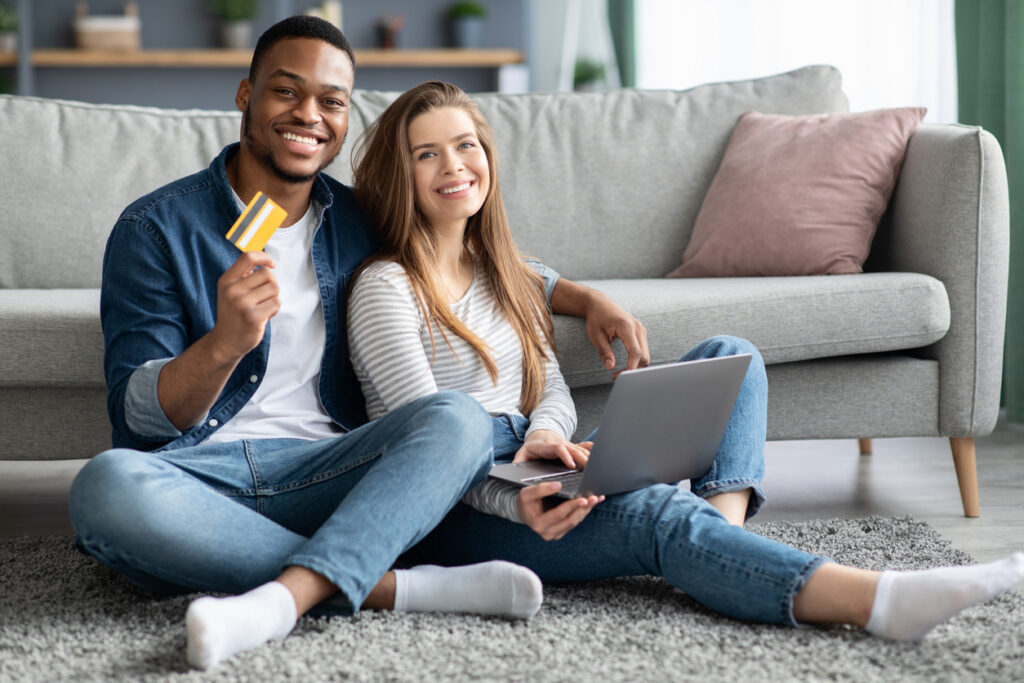Retrato de casal multicultural alegre com laptop e cartão de crédito avaliando quanto valem as milhas