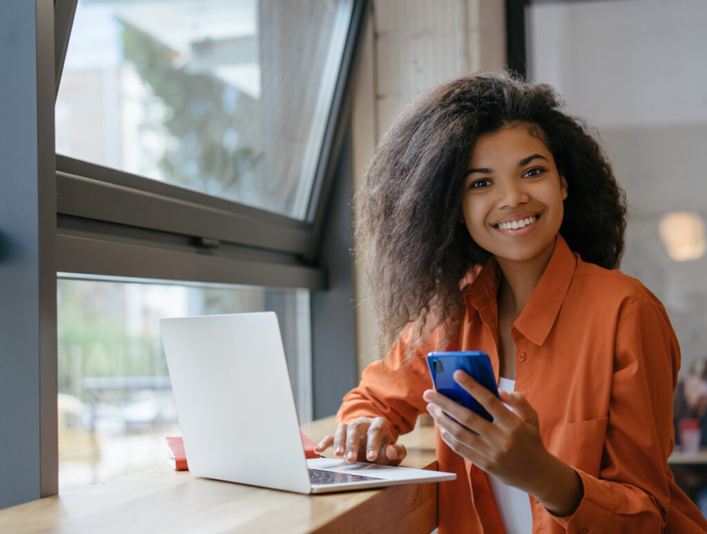 Linda mulher trabalhando no escritório. Retrato de empresária afro-americana sorridente usando computador portátil e internet, segurando o celular avaliando se o clube Smiles é bom para assinar.