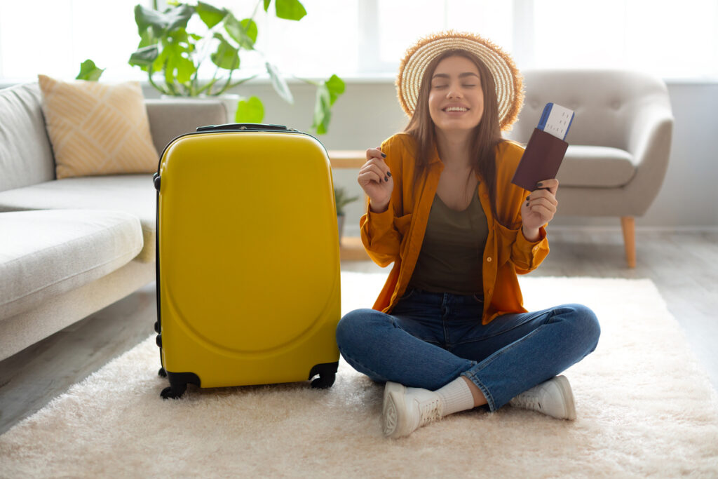 Nova viagem, finalmente. Animado jovem turista feminino segurando bilhete de viagem e passaporte, ansioso para o início das férias, sentado com mala no chão em casa. Companhia aérea escolhida foi de acordo com seu perfil.
