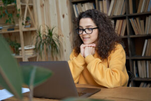 Uma menina de cabelo cacheado usando óculos e um moletom amarelo está sentada em uma mesa olhando para a tela do computador enquanto aprende sobre como funciona o processo de comprar ou vender milhas